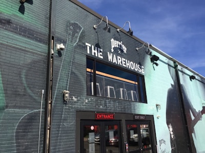 A brick building painted with a large mural featuring musical instruments and the silhouette of a person playing a guitar. The building has a sign reading 'The Warehouse' with the Perrier logo above it. There are industrial lights mounted on the wall and a glass entrance indicating entry and exit doors.