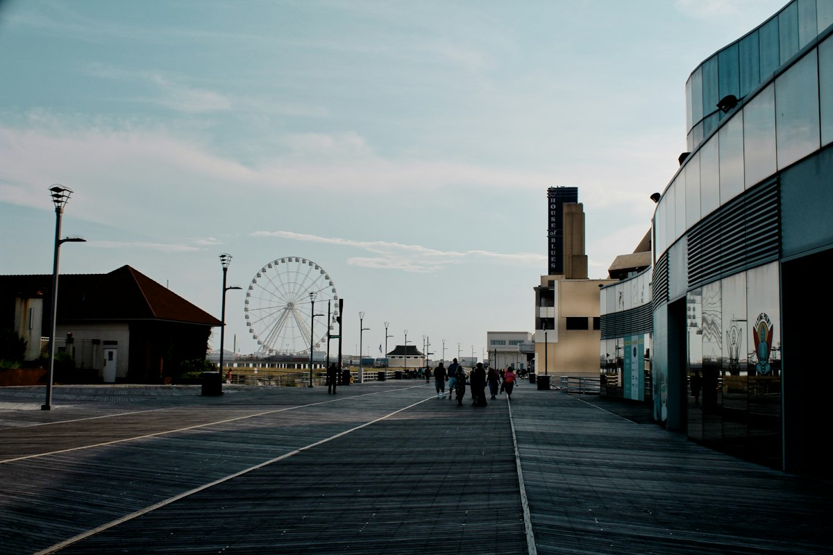 The Atlantic City Boardwalk running away into the distance, wide grey timber planking underfoot, a white Ferris wheel on a pier ahead, a tall tower carrying a vertical illuminated sign on the right, a curved glass building at the right edge, lamp posts and a few people walking further on