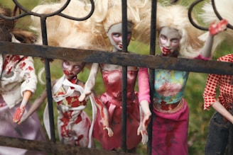 A group of eerie blonde dolls with disheveled hair and smeared with red paint resembling blood stand behind an iron fence, wearing tattered and stained clothing. The setting is outdoors with a grassy background.