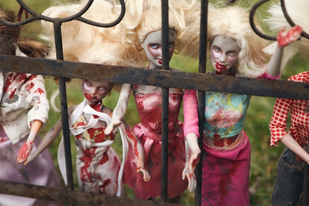 A group of eerie blonde dolls with disheveled hair and smeared with red paint resembling blood stand behind an iron fence, wearing tattered and stained clothing. The setting is outdoors with a grassy background.