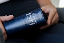A person is holding a blue leather-bound book labeled 'Academy Study Bible'. The book is grasped gently with the fingers visible, indicating a careful and contemplative handling.