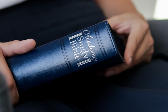 A person is holding a blue leather-bound book labeled 'Academy Study Bible'. The book is grasped gently with the fingers visible, indicating a careful and contemplative handling.