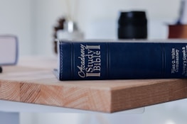 A blue Academy Study Bible with gold lettering is placed on a wooden surface. In the background, there are blurred objects including a white vase with dark stems and a black cylindrical container.