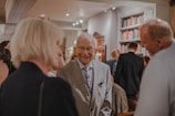 An elegant gentleman in a gray suit with a floral tie is smiling while engaging in conversation with a group of people. The setting appears to be a formal gathering or event inside a room decorated with book-filled shelves. The atmosphere seems lively with several people mingling in the background.