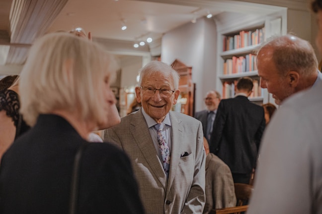 An elegant gentleman in a gray suit with a floral tie is smiling while engaging in conversation with a group of people. The setting appears to be a formal gathering or event inside a room decorated with book-filled shelves. The atmosphere seems lively with several people mingling in the background.