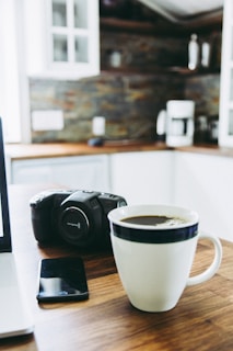 A cozy kitchen scene featuring a wooden table with a white coffee mug containing a dark beverage. A professional camera is placed next to a smartphone on the table. White cabinetry and a coffee maker are visible in the background, enhancing the warm and inviting ambiance.