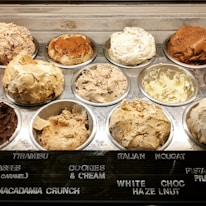 Various flavors of ice cream are displayed in a chilled dessert counter, each in its own metal tub with a different texture and color indicating the variety of flavors available. The names of the flavors such as tiramisu, cookies and cream, Italian nougat, and more are labeled on a board in front of the tubs.