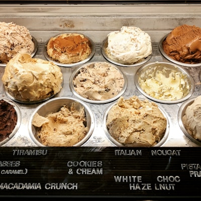Various flavors of ice cream are displayed in a chilled dessert counter, each in its own metal tub with a different texture and color indicating the variety of flavors available. The names of the flavors such as tiramisu, cookies and cream, Italian nougat, and more are labeled on a board in front of the tubs.
