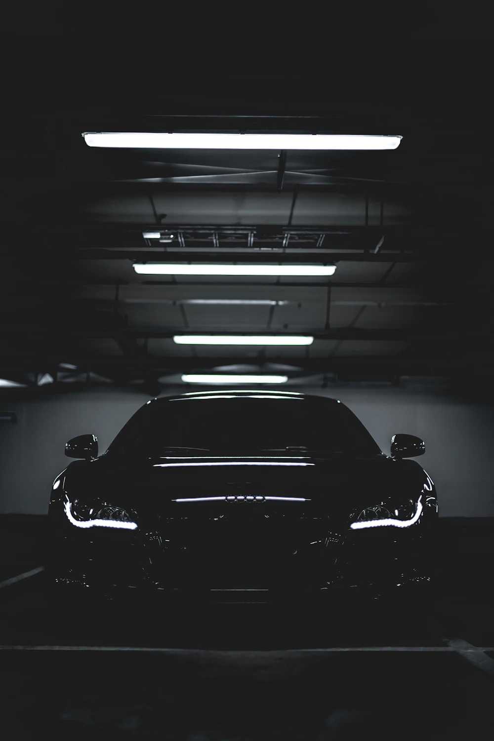 A dark coupe parked alone under fluorescent strip lights in an empty parking deck.