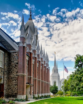 A grand brick building with intricate architecture and pointed spires is aligned next to a white temple with a golden figure atop its spire. A well-maintained lawn and garden are visible along with a prominently displayed American flag. Fluffy clouds fill the blue sky, adding to the sense of open space.