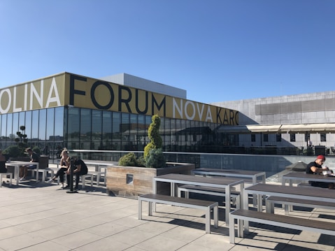 Outdoor seating area with several people sitting at benches on a spacious terrace. A modern building with large glass windows and a prominent sign reads 'FORUM'. The sky is clear and blue, and there are some decorative plants in planters.