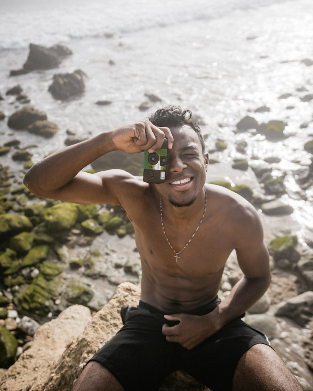 man wearing silver-colored cross necklace, and black shorts holding camera during daytime