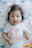 A baby is lying on a patterned blanket with a playful expression, sticking out their tongue. They have dark hair adorned with a white hair clip. The baby is wearing a white shirt with the phrase 'One of a Kind' printed in pink letters.