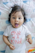 A baby is lying on a patterned blanket with a playful expression, sticking out their tongue. They have dark hair adorned with a white hair clip. The baby is wearing a white shirt with the phrase 'One of a Kind' printed in pink letters.