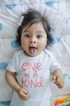 A baby is lying on a patterned blanket with a playful expression, sticking out their tongue. They have dark hair adorned with a white hair clip. The baby is wearing a white shirt with the phrase 'One of a Kind' printed in pink letters.
