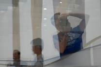 A reflection of a person holding a camera is visible on a glass surface, with their silhouette clearly outlined. In the background, there are blurred figures of other people and abstract geometric shapes, possibly pillars or walls, contributing to a layered composition.