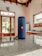 A blue portable speaker with a red logo sits on a polished kitchen countertop. The interior space includes wooden cabinetry, large windows letting in natural light, and visible appliances like a fridge. There is also a can of soda in the background.