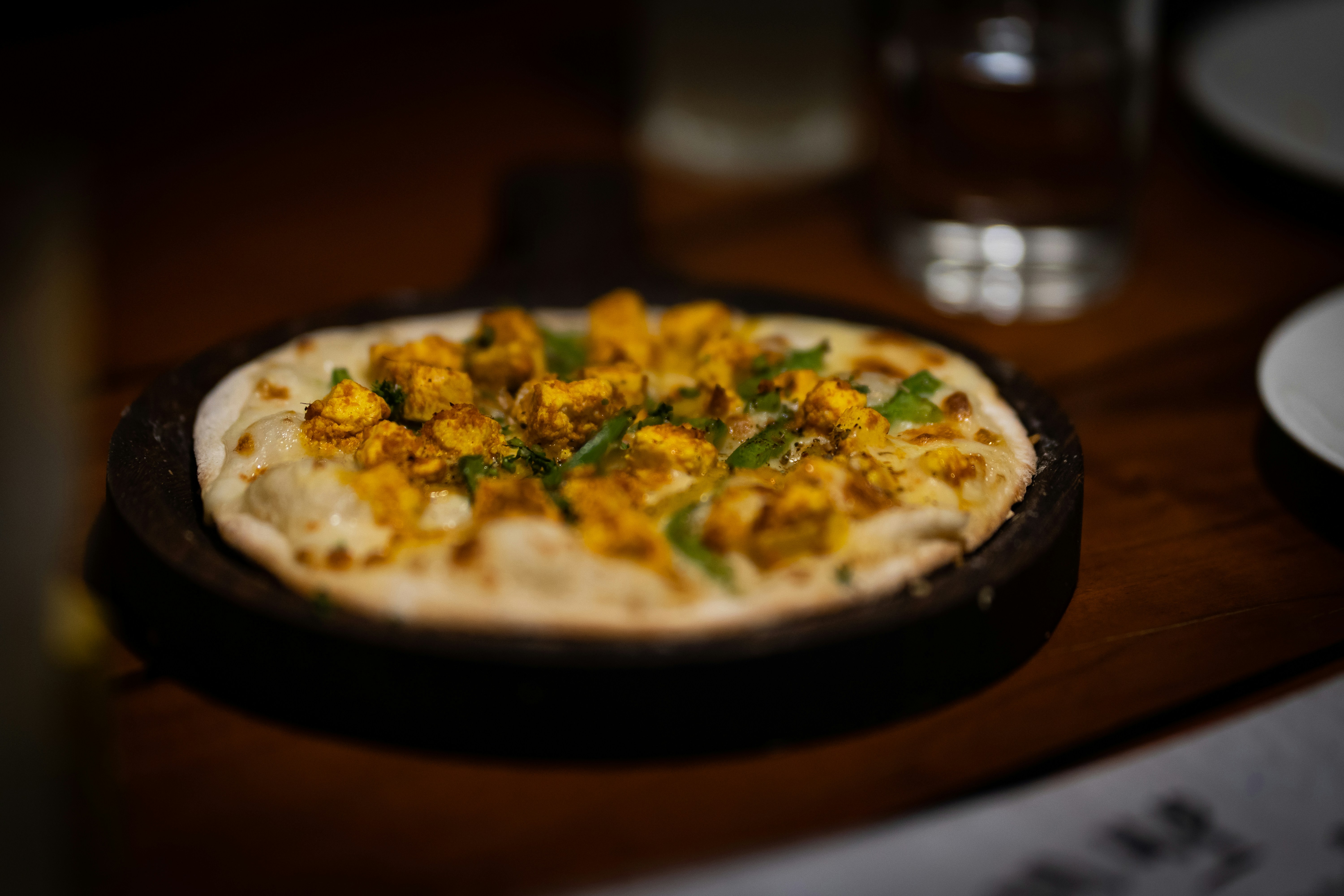 Rustic hand-made pizza with bubbling mozzarella and golden paneer on dark stone surface, warm oven-glow lighting