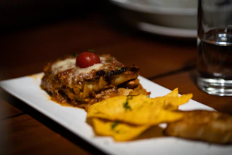 A delicious serving of lasagna topped with melted cheese and a small cherry tomato is placed on a white rectangular plate. Next to the lasagna are several crispy tortilla chips garnished with herbs. A glass of water and some crockery can be seen in the background, all set on a wooden table.