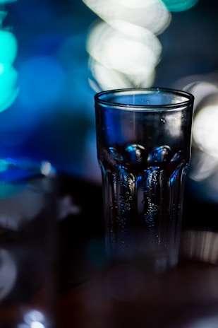 A clear glass with condensation droplets on its surface, set against a blurred background with bokeh lights in hues of blue and white. The lighting creates a reflective effect on the glass.