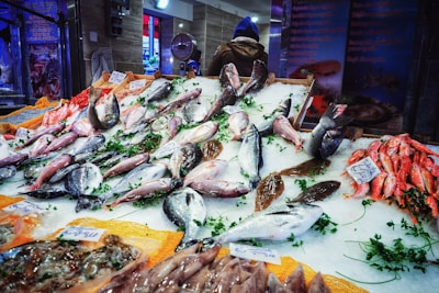A bustling fish market stall with a variety of fresh fish displayed on ice. Different species, including larger fish in the center and smaller red fish to the right, are arranged neatly, some labeled with price tags. Garnished with green herbs, the display is vibrant and colorful. A person in a blue hoodie appears in the background, facing away.
