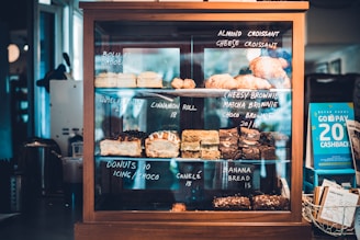 A wooden and glass display case filled with assorted pastries and baked goods, each with handwritten labels indicating their names and prices. Items include almond croissants, cheesy brownies, cinnamon rolls, and banana bread. A promotional card to the right offers a cashback deal.