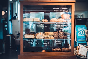 A wooden and glass display case filled with assorted pastries and baked goods, each with handwritten labels indicating their names and prices. Items include almond croissants, cheesy brownies, cinnamon rolls, and banana bread. A promotional card to the right offers a cashback deal.