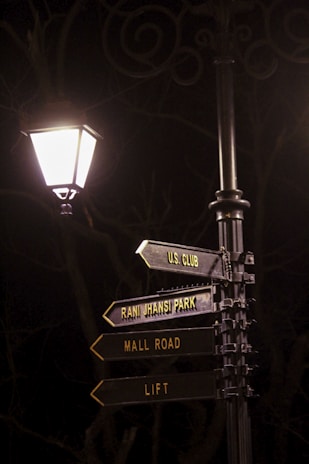 A street lamp illuminates a metal signpost at night. The signpost has several directional signs pointing to locations such as 'U.S. Club', 'Rani Jhansi Park', 'Mall Road', and 'Lift'. The background is dimly lit, with shadows and faint outlines of trees.