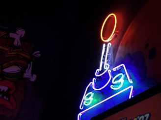 A neon sign glows vibrantly against a dark background, featuring a design that looks like a tank with a ball or sun above it. The neon lights emit colors such as red, blue, and green, creating an eye-catching visual contrast. To the left, a mural or painting of a cartoonish face with exaggerated features, including a prominent nose and mouth, is partially visible.