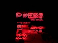 A glowing neon sign in bold red with the words 'PRESS PLAY' displayed prominently. The background is dark, which enhances the brightness and attention of the red neon letters and arrows pointing to the right.