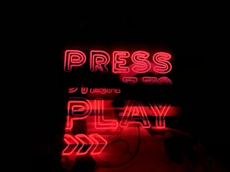 A glowing neon sign in bold red with the words 'PRESS PLAY' displayed prominently. The background is dark, which enhances the brightness and attention of the red neon letters and arrows pointing to the right.