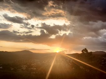 A scenic sunset view over a hilly landscape with scattered dark clouds. The sun is partially hidden behind the hills, casting a warm golden glow across the sky. The landscape features several trees and what appears to be a small town in the distance.