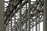 A complex metallic structure with numerous intersecting beams and rivets, creating a pattern of lines and angles. The beams display metalwork typical of industrial or infrastructure constructions.