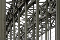 A complex metallic structure with numerous intersecting beams and rivets, creating a pattern of lines and angles. The beams display metalwork typical of industrial or infrastructure constructions.