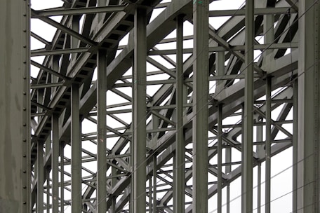 A complex metallic structure with numerous intersecting beams and rivets, creating a pattern of lines and angles. The beams display metalwork typical of industrial or infrastructure constructions.