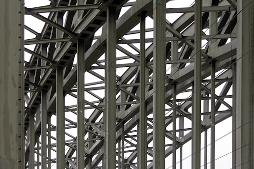 A complex metallic structure with numerous intersecting beams and rivets, creating a pattern of lines and angles. The beams display metalwork typical of industrial or infrastructure constructions.