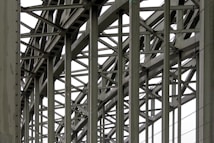 A complex metallic structure with numerous intersecting beams and rivets, creating a pattern of lines and angles. The beams display metalwork typical of industrial or infrastructure constructions.