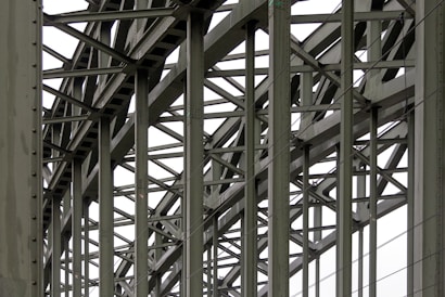 A complex metallic structure with numerous intersecting beams and rivets, creating a pattern of lines and angles. The beams display metalwork typical of industrial or infrastructure constructions.