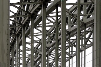 A complex metallic structure with numerous intersecting beams and rivets, creating a pattern of lines and angles. The beams display metalwork typical of industrial or infrastructure constructions.