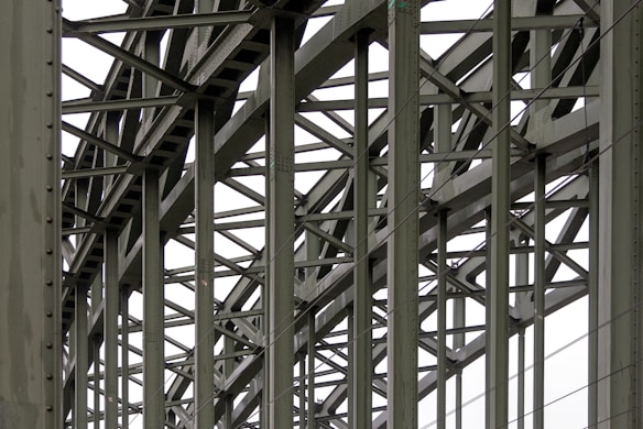 A complex metallic structure with numerous intersecting beams and rivets, creating a pattern of lines and angles. The beams display metalwork typical of industrial or infrastructure constructions.