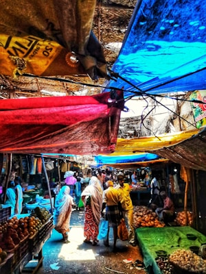 A bustling market scene with vibrant fabric canopies providing shade. People are walking and shopping, surrounded by various stalls displaying fruits and vegetables. The environment is lively and colorful, with sunlight filtering through colored tarps, casting multicolored shadows.