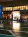 A nighttime urban street scene featuring a vape shop with bright neon lights, reflecting on the wet pavement. The storefront is illuminated, contrasting with the dim surroundings. A few people are visible near the shop, possibly customers or pedestrians, and a large blue vehicle is partially visible on the left.