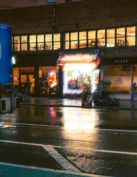 A nighttime urban street scene featuring a vape shop with bright neon lights, reflecting on the wet pavement. The storefront is illuminated, contrasting with the dim surroundings. A few people are visible near the shop, possibly customers or pedestrians, and a large blue vehicle is partially visible on the left.