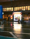 A nighttime urban street scene featuring a vape shop with bright neon lights, reflecting on the wet pavement. The storefront is illuminated, contrasting with the dim surroundings. A few people are visible near the shop, possibly customers or pedestrians, and a large blue vehicle is partially visible on the left.