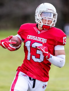 A football player wearing a red jersey with the number 13 and the word 'CRUSADERS' holds a football while running. He is equipped with a white helmet and red gloves, and the background appears to be an outdoor field.
