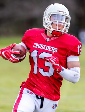 A football player wearing a red jersey with the number 13 and the word 'CRUSADERS' holds a football while running. He is equipped with a white helmet and red gloves, and the background appears to be an outdoor field.