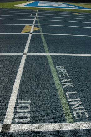 View of a running track with multiple lanes, white lane dividers, and a conspicuously marked break line. The number 100 is clearly visible along with a directional arrow pointing forward. A section of the track is highlighted in blue with large white lettering in the distance.