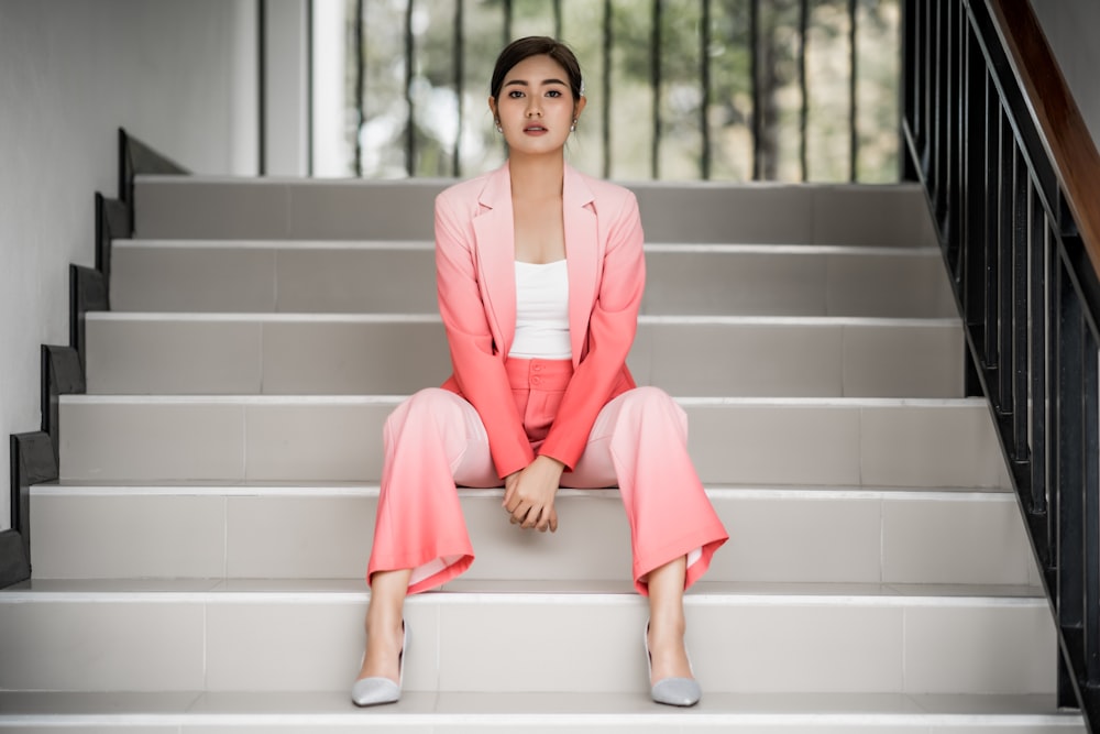 woman in pink blazer and pink pants sitting on white concrete steps indoors