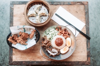A table setting featuring a variety of Asian dishes, including a stainless steel plate with rice topped with a reddish sauce, surrounded by peanuts, cucumber slices, what appears to be fried anchovies, half-boiled eggs, and some kind of meat. To the left, there is a basket of crispy fried chicken pieces lined with paper, and above it, a bamboo steamer containing dumplings. A pair of chopsticks rests on a white napkin on the wooden table surface.