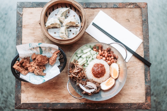A table setting featuring a variety of Asian dishes, including a stainless steel plate with rice topped with a reddish sauce, surrounded by peanuts, cucumber slices, what appears to be fried anchovies, half-boiled eggs, and some kind of meat. To the left, there is a basket of crispy fried chicken pieces lined with paper, and above it, a bamboo steamer containing dumplings. A pair of chopsticks rests on a white napkin on the wooden table surface.